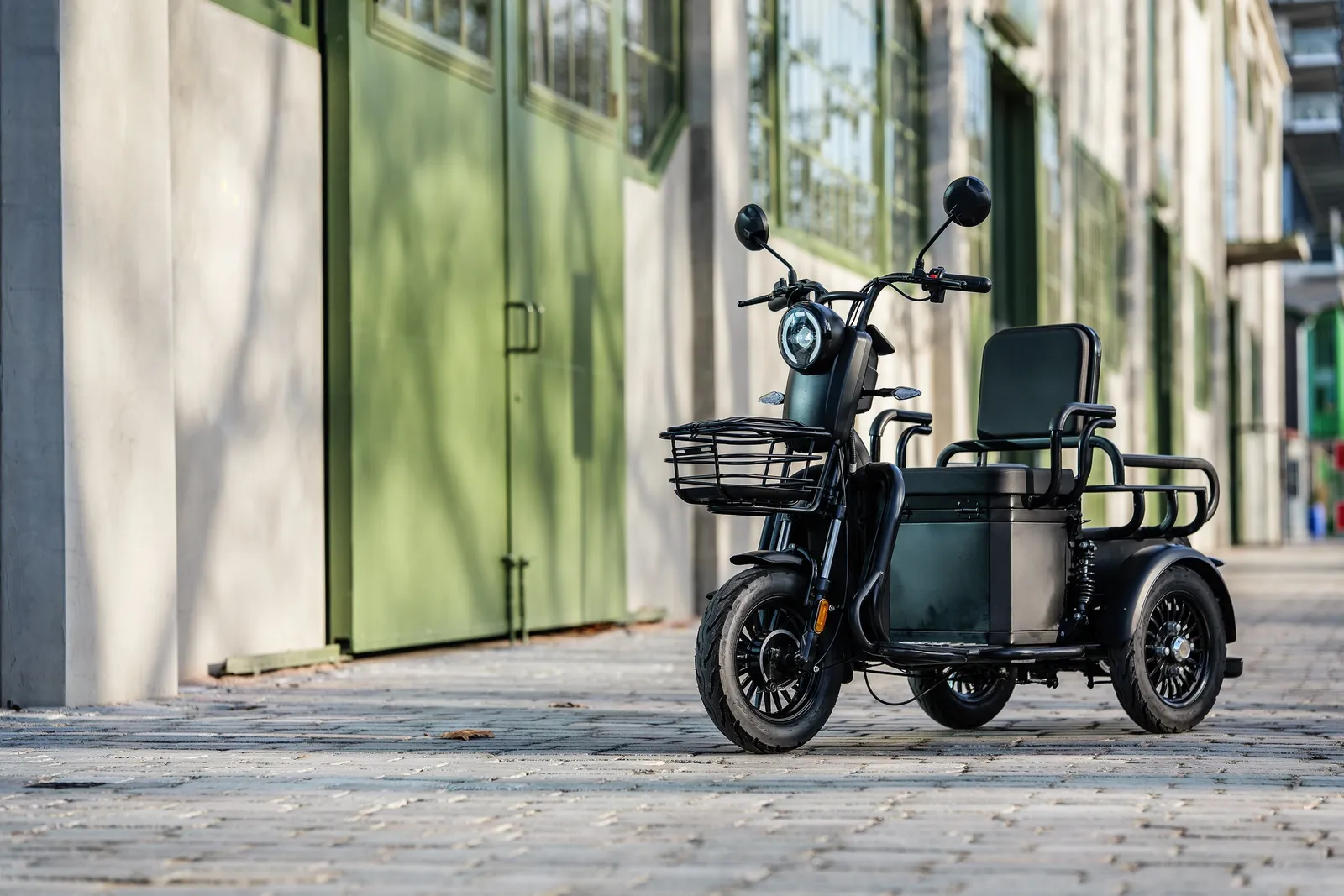 Een zwarte For Motion Cargos scootmobiel met voormand en bijrijdersbank in een steegje langs groene industriële deuren