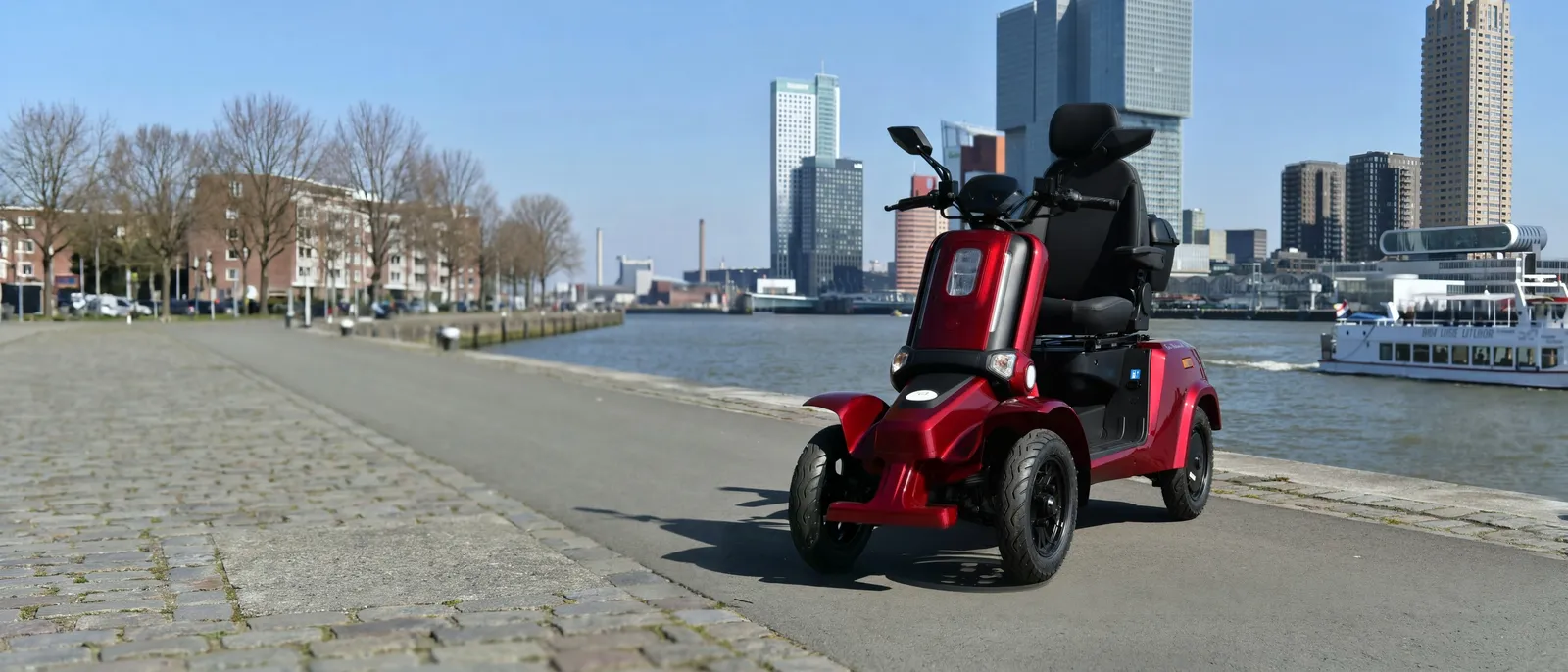 Een rode For Motion Forty-5 scootmobiel op de kade aan de Maas in Rotterdam, met de skyline van de Kop van Zuid en een passerende boot op de achtergrond