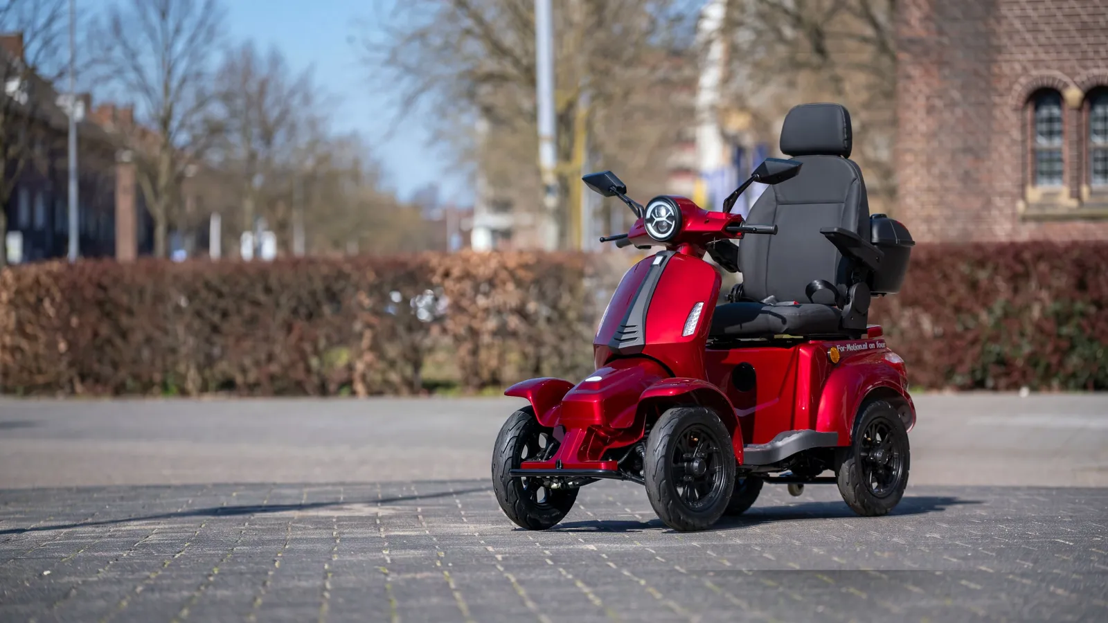 Een rode For Motion On Four scootmobiel schuin van voren op een straat voor een bakstenen kerk